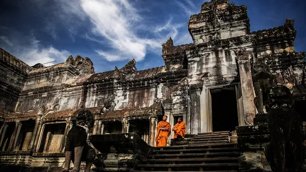 Dwóch mnichów w pomarańczowych szatach na stromych schodach galerii Angkor Wat, nad nimi błękitne niebo