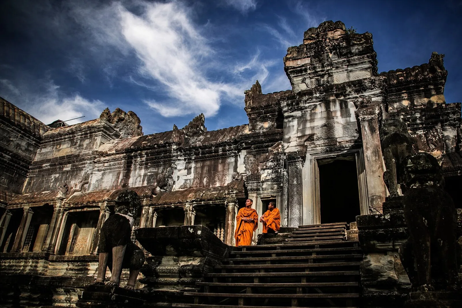 Dwóch mnichów w pomarańczowych szatach na stromych schodach galerii Angkor Wat, nad nimi błękitne niebo