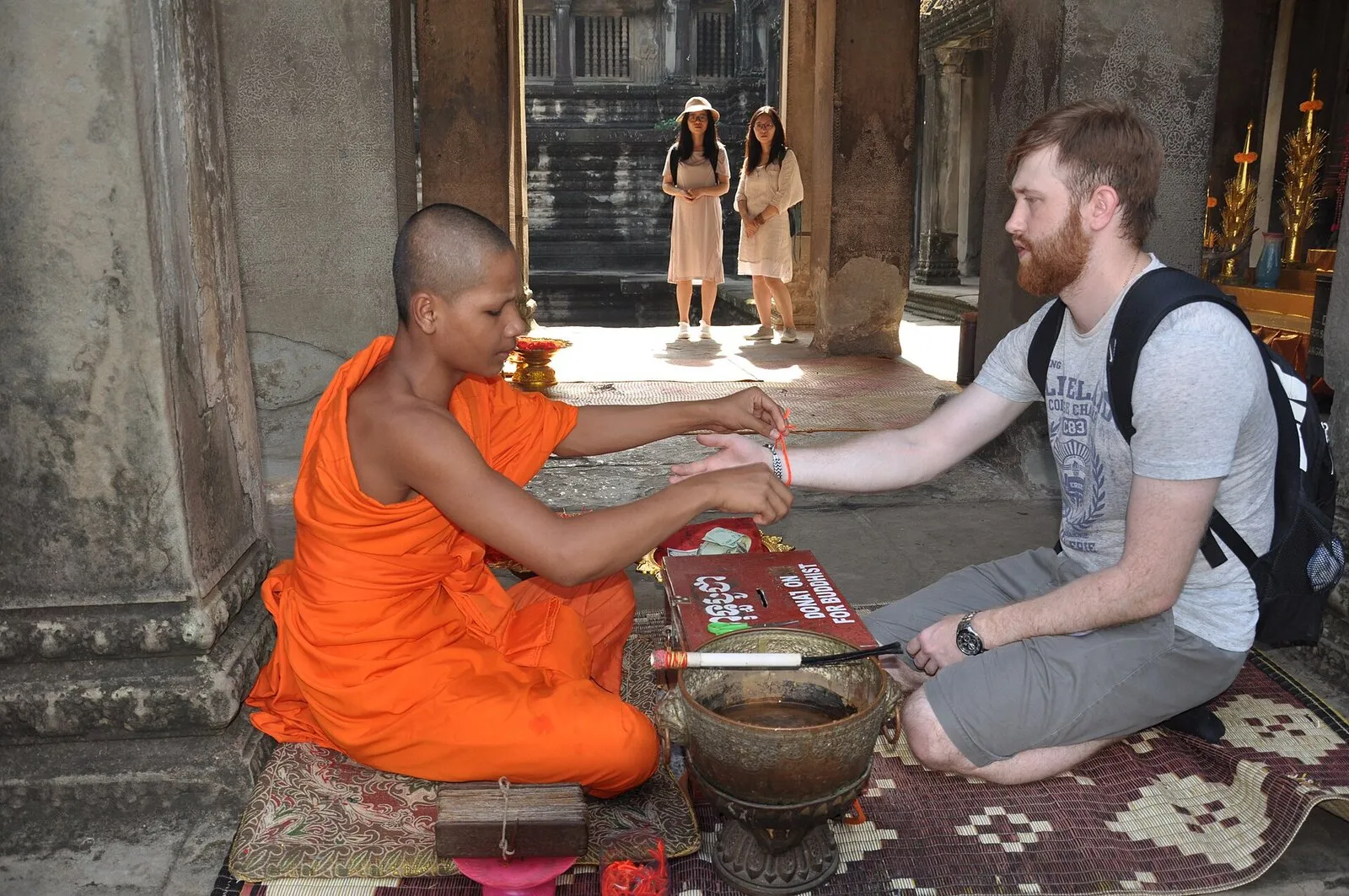 Mnich buddyjski w pomarańczowej szacie siedzi na macie i zawiązuje turyście na nadgarstku błogosławioną nitkę w galerii Angkor Wat