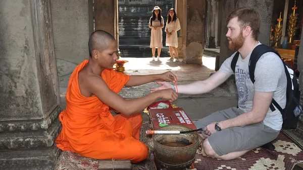 Mnich buddyjski w pomarańczowej szacie siedzi na macie i zawiązuje turyście na nadgarstku błogosławioną nitkę w galerii Angkor Wat