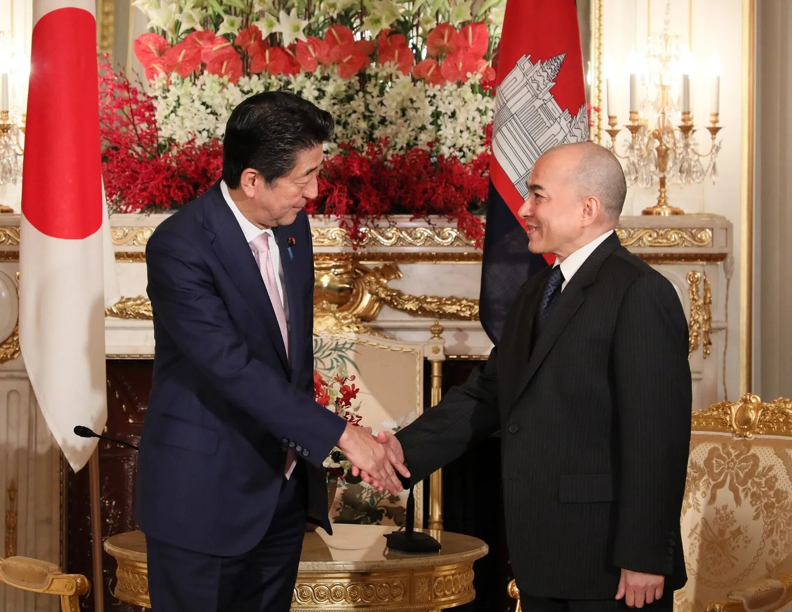 Król Norodom Sihamoni w ciemnym garniturze ściska dłoń premiera Japonii Shinzō Abe na tle flag Japonii i Kambodży