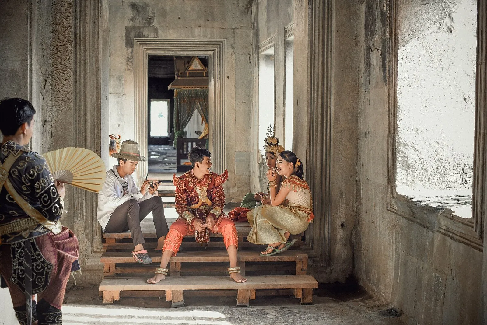 Para w tradycyjnych strojach khmerskich, mężczyzna w czerwonym sampocie chang kben, pozuje do zdjęcia w kamiennej galerii Angkor Wat, obok fotograf i osoba z wachlarzem