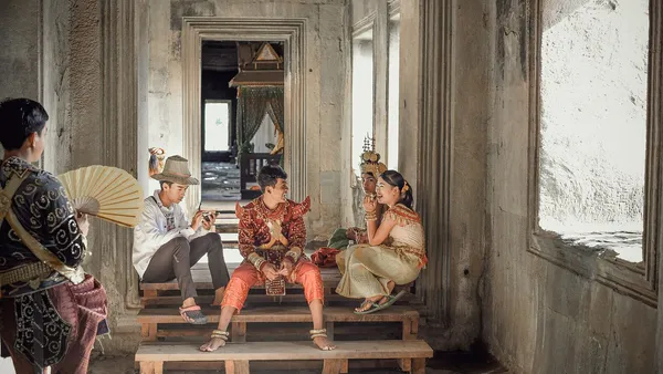 Para w tradycyjnych strojach khmerskich, mężczyzna w czerwonym sampocie chang kben, pozuje do zdjęcia w kamiennej galerii Angkor Wat, obok fotograf i osoba z wachlarzem