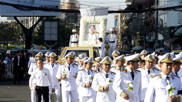 Kondukt pogrzebowy w Phnom Penh: portret zmarłego w ramie z białych kwiatów na karawanie, za nim urzędnicy w białych mundurach