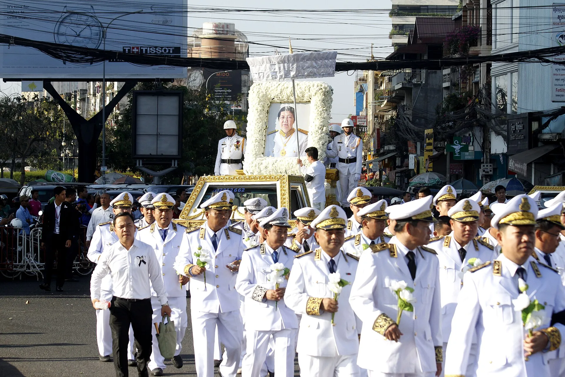 Kondukt pogrzebowy w Phnom Penh: portret zmarłego w ramie z białych kwiatów na karawanie, za nim urzędnicy w białych mundurach