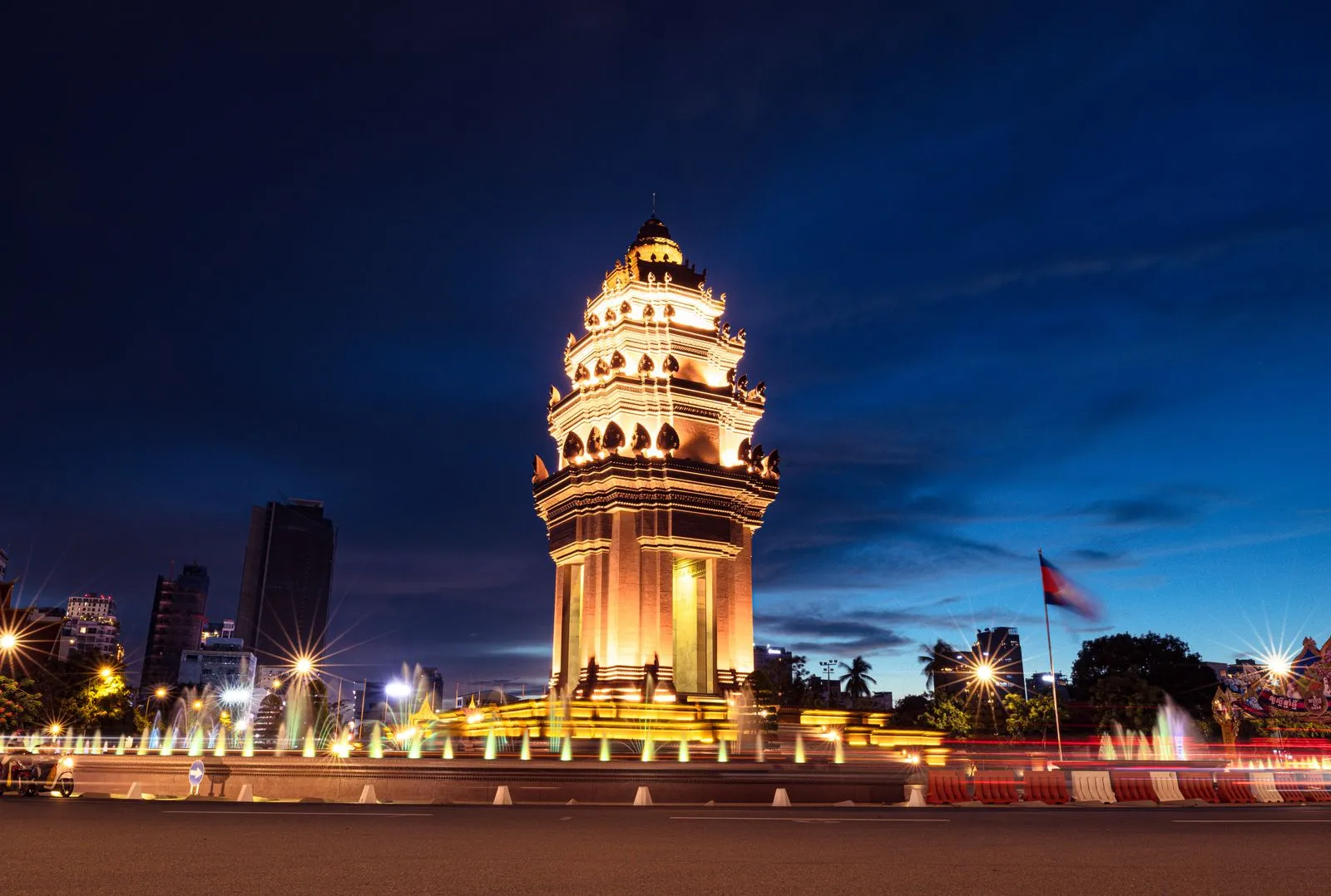 Podświetlony Pomnik Niepodległości w Phnom Penh po zmroku, obok fontanny i flaga Kambodży na maszcie